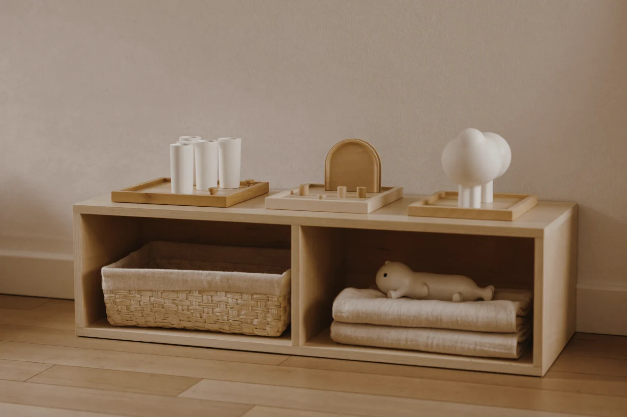 Une étagère basse en bois clair portant quatre activités Montessori sur leurs plateaux, un panier tressé et du linge plié en dessous