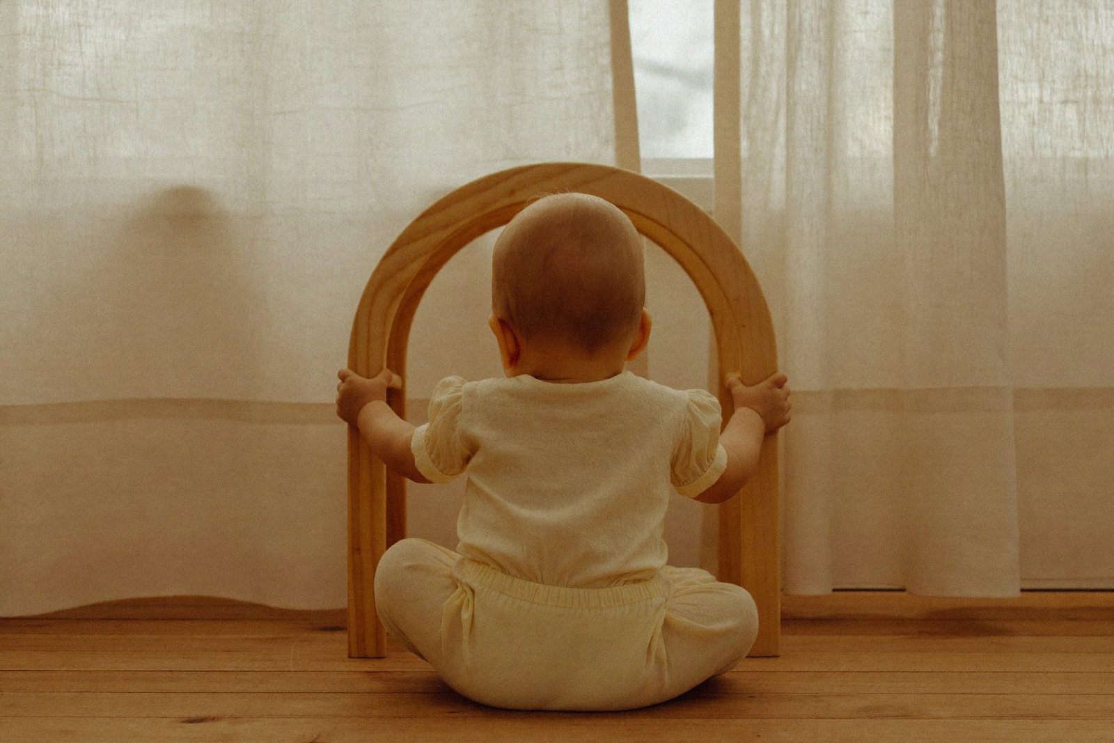 Bébé de dos jouant avec une arche à empiler en bois clair, dans une lumière douce de fin de journée
